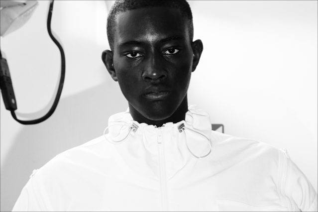 A male model snapped backstage in a white jacket at the Feng Chen Wang menswear show. Photographed by Alexander Thompson for Ponyboy magazine.